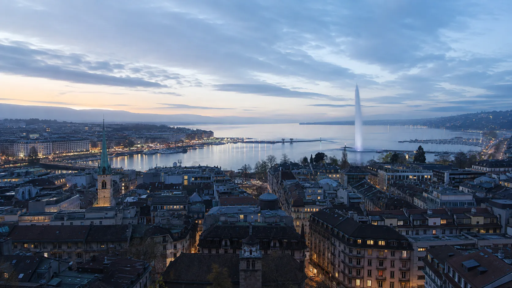 Genève au crépuscule vue depuis les hauteurs : les toits de la vieille ville, le lac, le jet d'eau et les lumières de la rade.